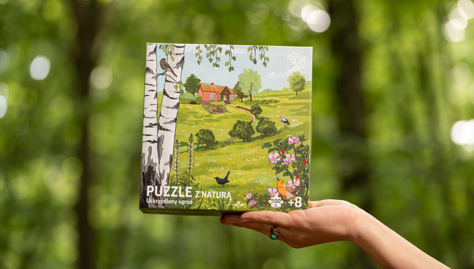 Puzzle z ogrodem, krajobrazem wsi, ptakami, kwiatami. Puzzle dla dorosłych, dla rodzin, nastolatków i seniorów. Na ilustracji rudzik, kos, pliszka siwa, kopciuszek zwyczajny, bocian biały oraz flora dzika róża, brzoza brodawkowata, naparstnica zwyczajna. W tle sad, dom tradycyjny, warzywny ogród i dzikie miejsca pozostawione dla owadow, mniejszych ssaków i ptaków.